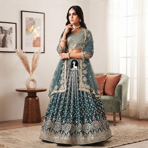 May include: A teal lehenga choli set featuring a sleeveless blouse and a floor-length skirt, both adorned with intricate gold embroidery and sequin detailing. A matching sheer teal dupatta with gold embroidered borders drapes over the shoulders.