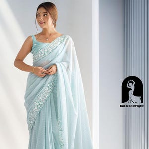 Może przedstawiać: Jasnoniebieskie sari z misternymi białymi i srebrnymi zdobieniami wzdłuż brzegów. Sari jest noszone na dopasowanej bluzce bez rękawów. Obraz zawiera logo "BOLD BOUTIQUE".