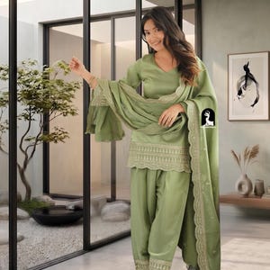 May include: A sage green three-piece salwar kameez set featuring a V-neck tunic, matching trousers, and a long dupatta. The tunic hem and trouser cuffs are adorned with intricate cream-colored embroidery and scalloped edges.