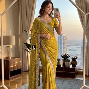 May include: A woman wearing a mustard yellow sari with silver sequin and lace trim. The sari has a matching blouse and is draped over the shoulder. The woman is holding a phone and is standing in front of a large window.