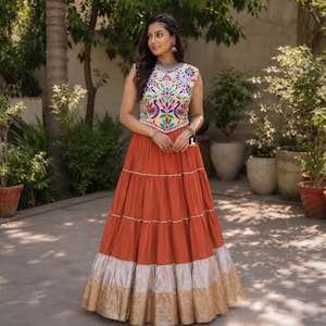 May include: A sleeveless floor-length dress featuring a white bodice with a colorful floral and peacock embroidery pattern. The skirt is a solid burnt orange color with horizontal metallic silver and gold bands at the hemline and thin silver trim.