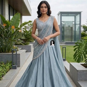 May include: A sleeveless slate blue lehenga choli featuring a fitted bodice with intricate silver embroidery and a matching pleated floor-length skirt. The ensemble includes a draped dupatta with a decorative silver border and a small logo graphic.