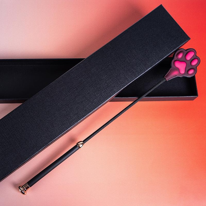 Op de afbeelding: Een zwarte flexibele zweep met een ros&eacute;gouden handvat en een decoratief uiteinde in de vorm van een pootafdruk. De pootafdruk heeft een donkerpaarse rand met een felroze centrum. Het item wordt gepresenteerd in een zwarte geschenkdoos.