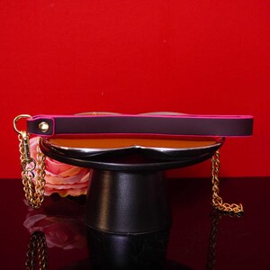 Pu&ograve; includere: Una borsa nera con una tracolla a catena dorata e un bordo rosa. La borsa &egrave; esposta su un supporto nero su sfondo rosso. La borsa ha una chiusura dorata e una catena dorata.