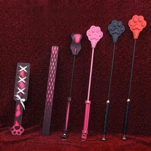 Op de afbeelding: Een verzameling van zes items, waaronder een roze en zwarte peddel met witte veters, een lange zwart-roze riem met patroon en vier dunne staven met pootvormige uiteinden in zwart, roze, donkergrijs en rood, allemaal voorzien van zwarte handvatten.