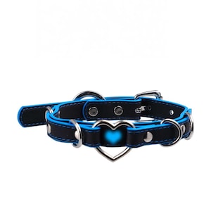 Könnte beinhalten: Ein schwarz-blaues Halsband mit einem herzförmigen Anhänger. Das Halsband hat eine blaue Umrandung und silberfarbene Metallbeschläge. Der Herzanhänger hat eine leuchtend blaue Mitte.