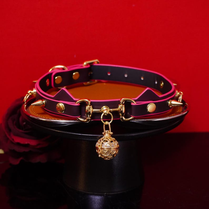 Pu&ograve; includere: Un collare nero e rosa con elementi dorati. Il collare presenta un design a orecchie di gatto, borchie appuntite e una campanella dorata pendente. Il collare &egrave; esposto su un supporto nero su sfondo rosso.