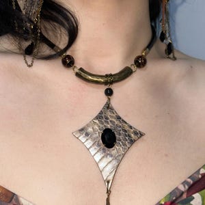 May include: A distinctive necklace and earring set. The necklace features a diamond-shaped pendant with a black stone and a snakeskin pattern. The necklace has a black cord with brass accents and dark brown beads. The earrings have a matching design with dangling black teardrop beads.