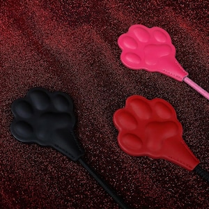 Op de afbeelding: Drie pootvormige voorwerpen in zwart, rood en roze, elk met een glad, gevormd oppervlak met duidelijke teenkussentjes en een centraal kussentje, bevestigd aan een dun, flexibel koord dat uit de basis van elke pootvormige kop komt.