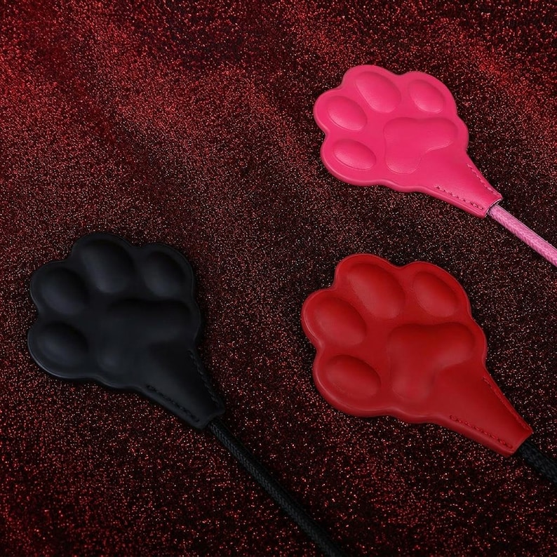 Op de afbeelding: Drie pootvormige voorwerpen in zwart, rood en roze, elk met een glad, gevormd oppervlak met duidelijke teenkussentjes en een centraal kussentje, bevestigd aan een dun, flexibel koord dat uit de basis van elke pootvormige kop komt.