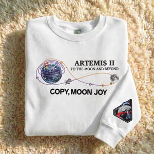 Puede incluir: Sudadera blanca de cuello redondo con un diseño gráfico que representa la Tierra, una nave espacial y la luna, con el texto "ARTEMIS II TO THE MOON AND BEYOND" y "COPY, MOON JOY". Un parche con un cohete y la luna está en la manga.