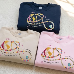 Puede incluir: Tres sudaderas en azul marino, beige y rosa, cada una con un diseño bordado. El diseño incluye un globo terráqueo, una pulsera de cuentas y el texto "COPY HEART BRACELET ARTEMIS". Las sudaderas tienen el texto "It's been a long time coming..."