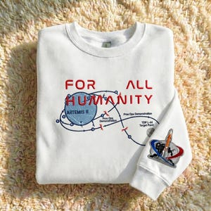 Puede incluir: Sudadera blanca con el texto rojo "FOR ALL HUMANITY". Un gráfico azul y blanco representa una misión espacial con "ARTEMIS II". La manga tiene un parche de cohete con "TDP L-44 Target Point".