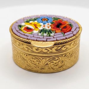 May include: A round gold-tone metal trinket box featuring an intricate floral micro-mosaic inlay on the lid. The lid is bordered by a light purple mosaic tile frame, while the sides of the box are decorated with an embossed floral scroll pattern.