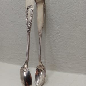 May include: A pair of silver-toned metal sugar tongs with a polished finish. The handle features an ornate, embossed oval design with scrollwork details. The spoon-shaped ends are smooth and rounded, and the inner arm displays small stamped hallmarks.