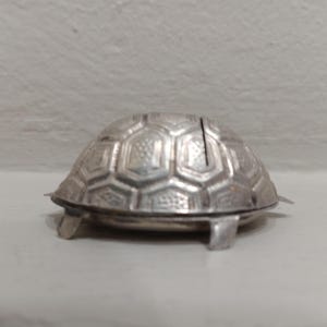 Könnte beinhalten: Eine silberfarbene Metalldose in Schildkrötenform mit aufklappbarem Panzerdeckel. Der Panzer weist ein erhabenes Sechseckmuster mit texturierten Details auf jedem Segment auf. Das Objekt hat vier kleine Metallfüße und eine glatte Basis.