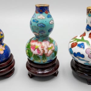 Pode incluir: Três vasos cloisonné de várias formas e tamanhos, cada um com padrões intrincados de flores e nuvens em esmalte azul, branco, rosa e verde. Cada vaso é apoiado por um suporte de exibição circular de madeira castanho-escura com pés.