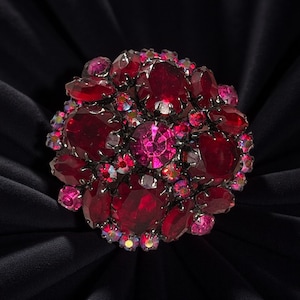 Op de afbeelding: Een ronde broche met rode en roze edelstenen. De broche heeft grote, ovale rode stenen omringd door kleinere, roze en iriserende kristallen. De zetting is van donker metaal, mogelijk zilver, tegen een zwarte achtergrond.