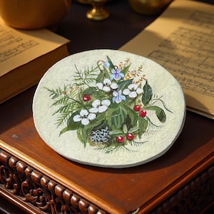 May include: A round, off-white painted coaster with a floral design. It features white flowers, red berries, green leaves, and blue accents. The coaster is placed on a dark wooden surface.