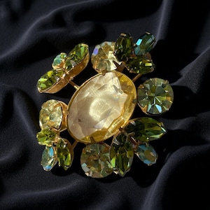 Puede incluir: Broche dorado con una piedra central ovalada de color amarillo claro, rodeada de cristales verdes y verde amarillento de varios tamaños. El broche tiene un diseño floral, con los cristales dispuestos para parecer hojas y pétalos.