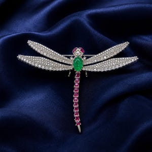 May include: A dragonfly brooch crafted in silver-toned metal, featuring wings encrusted with clear white stones. The body displays a vibrant green oval center stone, a long tail set with square-cut pink stones, and two small red eyes.