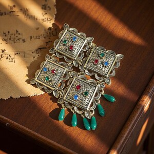 May include: A vintage gold-coloured brooch featuring four square panels, each set with red, green, and blue gemstones. The brooch has a floral design around the squares and green teardrop-shaped beads hanging from the bottom.