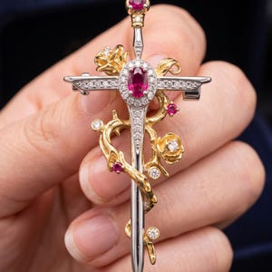 Könnte beinhalten: Eine schwertförmige Brosche aus silber- und goldfarbenem Metall. Das Heft zeigt einen zentralen ovalen Rubin, umgeben von kleinen weißen Steinen. Eine goldene Ranke mit modellierten Rosen sowie kleinen Rubinen und weißen Steinen umwindet die Klinge.