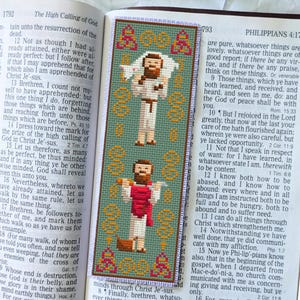 May include: A green and gold cross-stitch bookmark featuring two figures with brown hair and beards, one holding a lamb. The bookmark is placed in an open book with text visible on both pages. The bookmark has a decorative border.