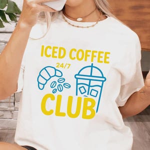 May include: Off-white t-shirt featuring the text "ICED COFFEE CLUB" in yellow and blue. The design includes a croissant, coffee beans, and a takeaway coffee cup. A casual, everyday wear item.