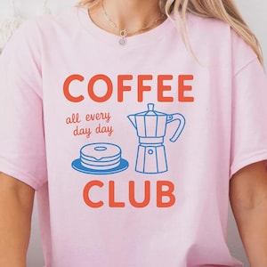 May include: A light pink t-shirt with the words "COFFEE CLUB" in orange, and "all every day day" in a smaller font. A blue illustration of a coffee maker and a stack of pancakes is between the words. The shirt is a casual style.