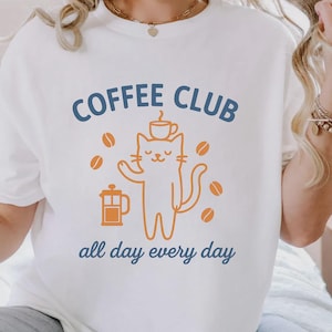 May include: White t-shirt with a blue and orange graphic. The graphic says "COFFEE CLUB" above a cat illustration holding a coffee cup, surrounded by coffee beans. Below the cat, the text reads "all day every day."