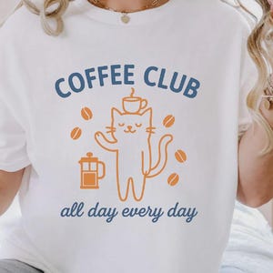 May include: White t-shirt with a blue and orange graphic. The design features the words "COFFEE CLUB" above a cat illustration holding a coffee cup, with coffee beans around it. The phrase "all day every day" is below the cat.