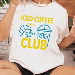 May include: Cream-colored t-shirt with the words "ICED COFFEE CLUB" in yellow and blue. The design includes a croissant, coffee beans, and a to-go coffee cup. The shirt is worn by a person.