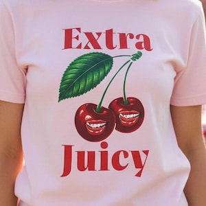 May include: A pale pink t-shirt featuring the text "Extra Juicy" in red, accompanied by a graphic of two cherries with smiling faces. The short-sleeved top is being worn outside.