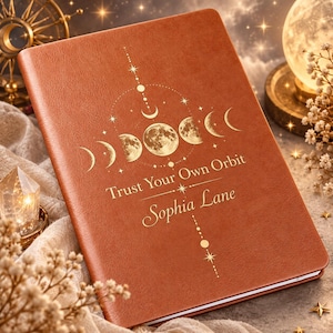 May include: A brown notebook featuring a gold foil moon phase graphic, the text "Trust Your Own Orbit," and the name "Sophia Lane" printed on the front cover. The design includes celestial stars and vertical line accents.