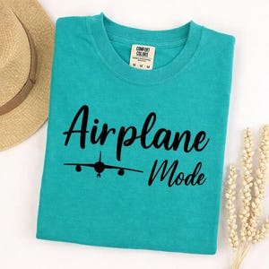 Sea Foam Comfort Colors tee folded with black Airplane Mode text and airplane icon, stylish travel shirt for vacation wear.