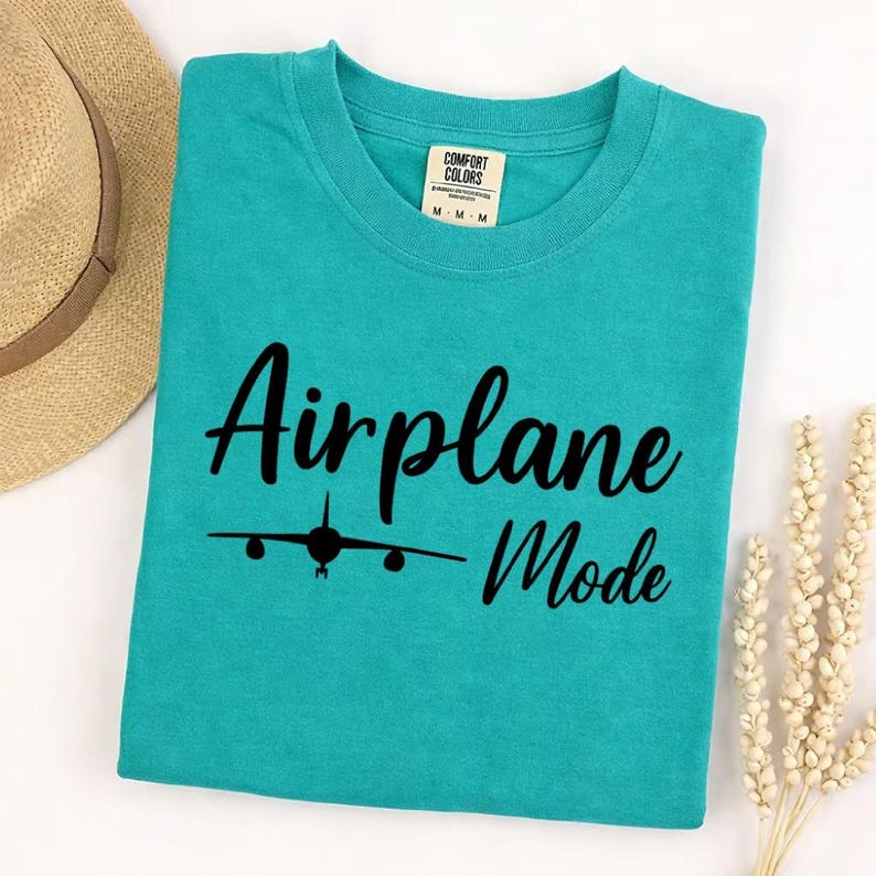 Sea Foam Comfort Colors tee folded with black Airplane Mode text and airplane icon, stylish travel shirt for vacation wear.