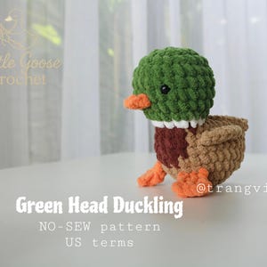 May include: A handmade crochet duckling with a green head, brown body, and orange beak and feet. The duckling has a white collar and black eyes. The text "Green Head Duckling" is below the duckling.