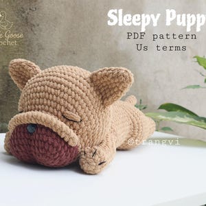 Könnte beinhalten: Ein gehäkelter Welpe in Schlafposition, mit hellbraunem Körper und dunklerer brauner Schnauze. Der Welpe hat geschlossene Augen und eine Knopf-Nase. Der Text "Sleepy Puppy PDF pattern Us terms" wird angezeigt.