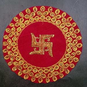 May include: A circular, red decorative piece with gold embroidery. The centre displays a gold embroidered Swastika symbol. The outer edge is decorated with gold floral patterns and sequins. This item is likely used for cultural or religious purposes.