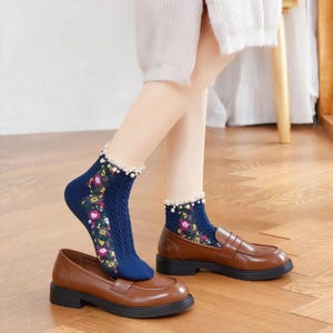 May include: A pair of navy blue ankle socks featuring a vertical cable knit pattern, a colorful floral print along the side, and a white ruffled lace trim at the cuff adorned with small white faux pearls, paired with brown leather loafers.