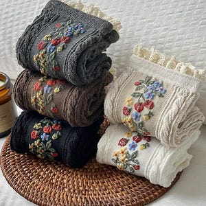 Könnte beinhalten: Ein Stapel aus fünf gefalteten Socken in Anthrazit, Braun, Schwarz und Creme. Jede Socke verfügt über ein strukturiertes Zopfstrickmuster, einen gerüschten Spitzenbesatz am Bündchen und ein gesticktes Blumenmuster mit roten, blauen und gelben Blüten.