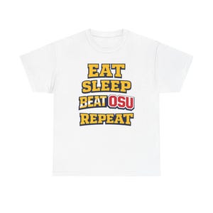 May include: White t-shirt with the text "EAT SLEEP BEAT OSU REPEAT" in yellow and blue block letters. The word "OSU" is in red. The shirt is a casual wear item.