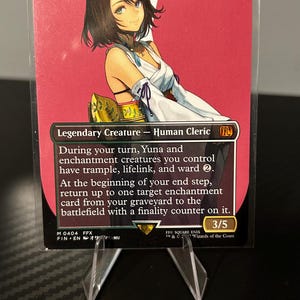 May include: A trading card featuring Yuna, Hope of Spira, a legendary Human Cleric. The card has a red background with an illustration of a woman. The card is displayed in a protective sleeve and held by a clear stand.