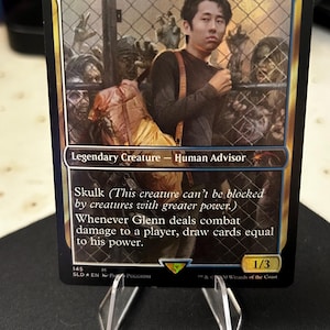 May include: A Magic: The Gathering card featuring "Glenn, the Voice of Calm." The card shows a man with a backpack, with zombies behind a fence. The card text includes "Legendary Creature - Human Advisor" and game instructions. The card is on a clear stand.