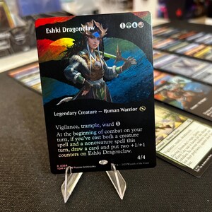 May include: A Magic: The Gathering card depicting Eshki Dragonclaw, a Legendary Creature Human Warrior. The card has a vibrant background and text detailing its abilities and stats. It is presented on a clear plastic stand.