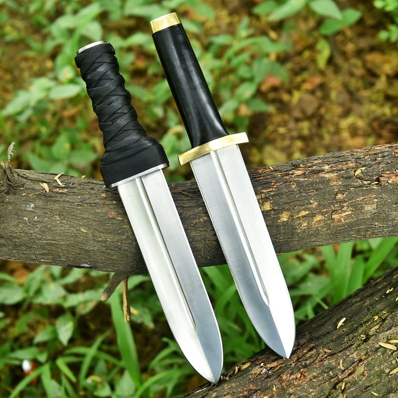 May include: Two silver-bladed daggers with black handles and gold accents rest on a wooden branch. One handle is wrapped in black leather. The blades are sharp and pointed, reflecting the light. The background is blurred green foliage.