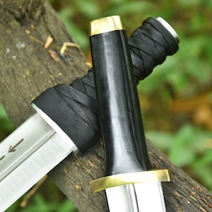 May include: Two swords with silver blades and black handles, with gold-coloured accents. The handles are wrapped in black leather. The swords are resting on a piece of wood, with a blurred green background.