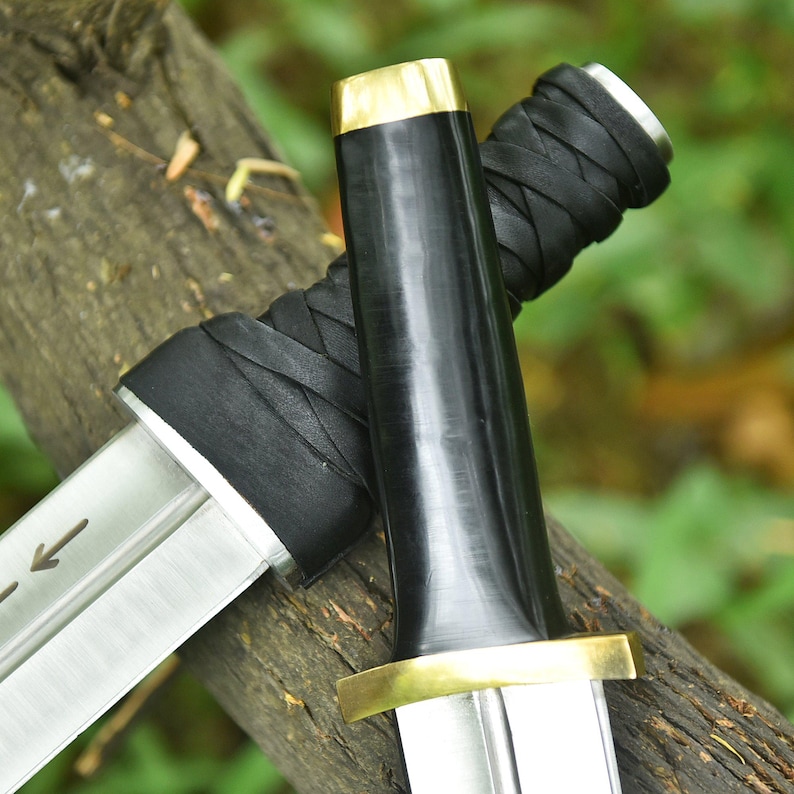 May include: Two swords with silver blades and black handles, with gold-coloured accents. The handles are wrapped in black leather. The swords are resting on a piece of wood, with a blurred green background.