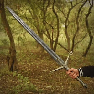 Peut inclure: Une longue épée argentée avec une garde et une poignée dorées ornées, sur un fond de forêt flou. La lame présente une finition texturée. La poignée comporte des motifs détaillés.